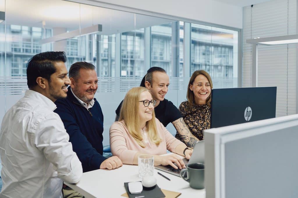 A group of people from Billwerk+ stand in front of a standing desk and look at the computer screen. Everyone is very happy. 