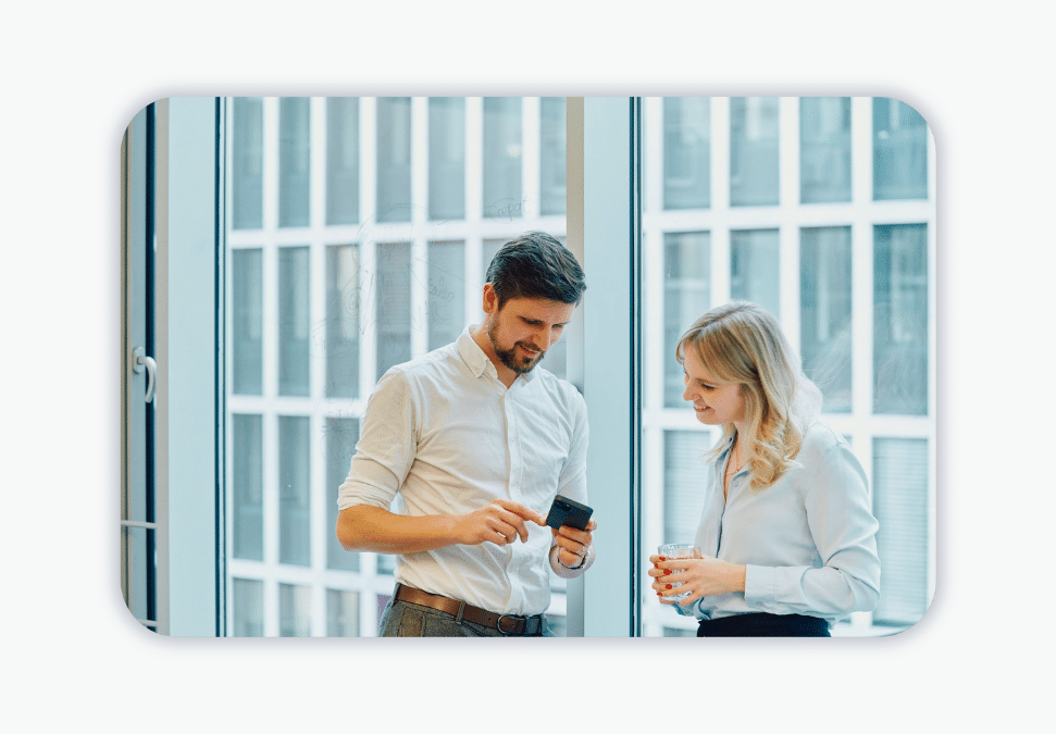 Two people stand at a large office window and look at a smartphone. 