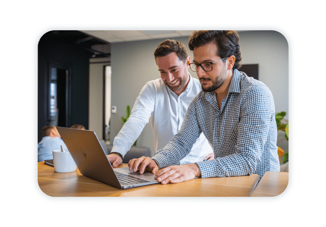 Two people are at a standing desk and look at a laptop.