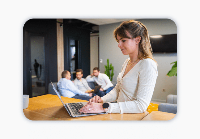 A woman works at a laptop at a standing desk. Three colleagues sit in the background and have a conversation