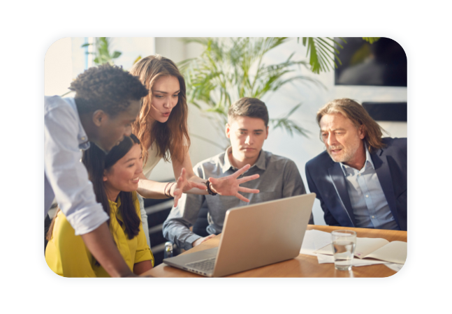 A group of colleagues sit together at a desk and discuss something they see on a laptop.